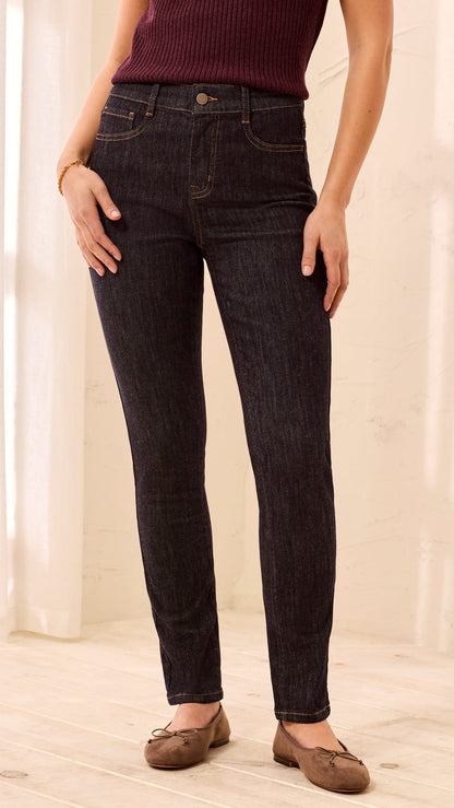 A person wearing dark indigo skinny jeans, a burgundy ribbed top, and brown flat shoes, standing in a brightly lit room with light-colored walls and wood flooring.