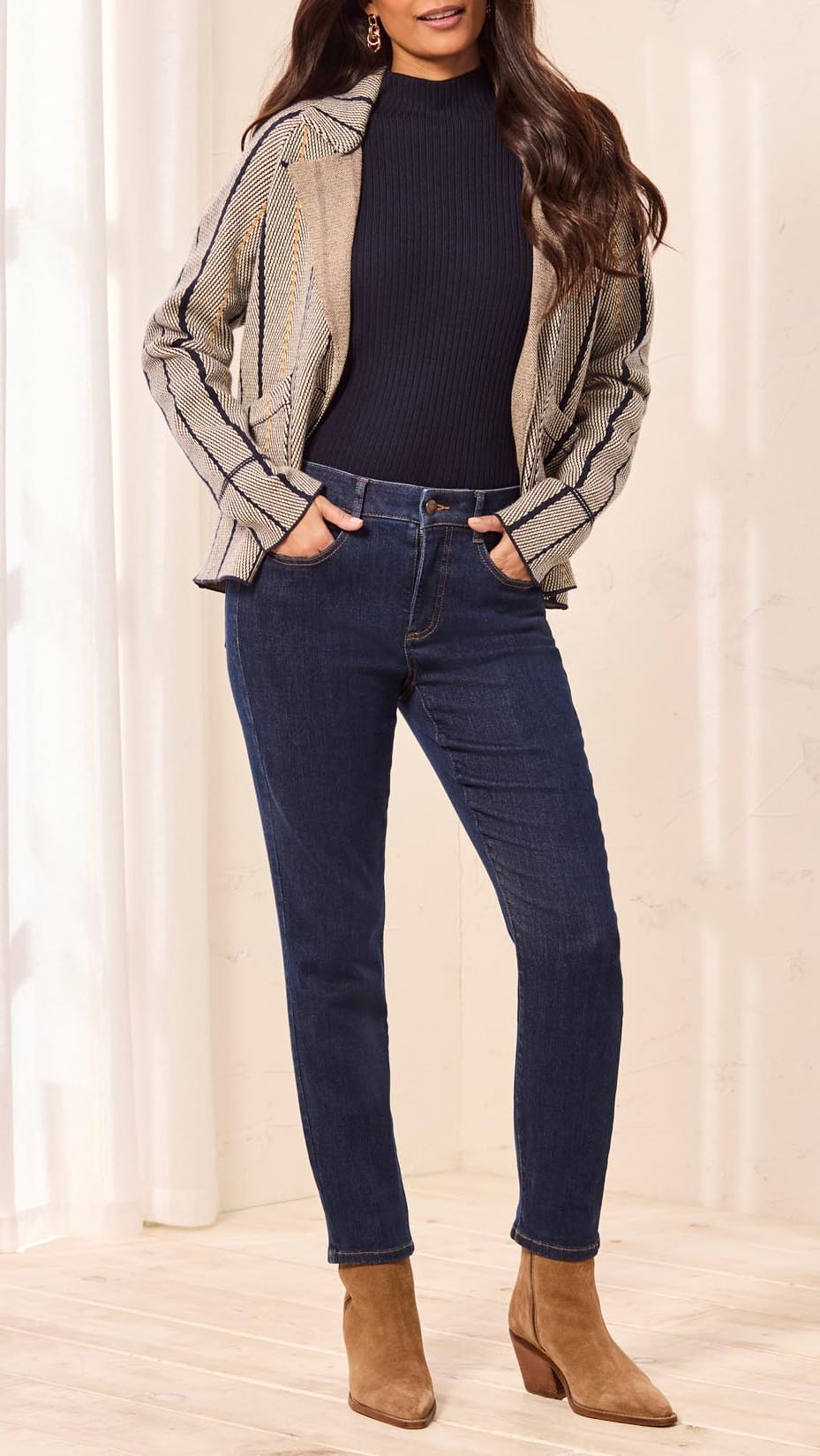 A woman modeling a beige and black striped moto jacket over a dark navy ribbed turtleneck, paired with dark blue skinny jeans and brown suede ankle boots, standing in a brightly lit indoor setting.