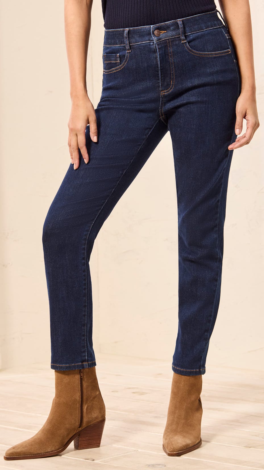 A model wearing dark blue slim-fit jeans and brown suede ankle boots, standing against a light beige wall and wood floor.