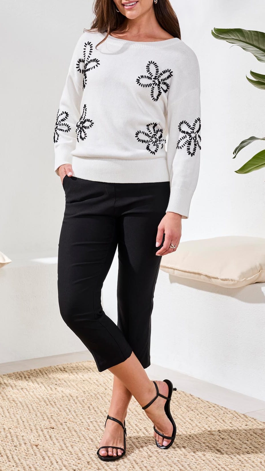 Woman wearing a white sweater with black floral embellishments and black pants in a minimal indoor setting.