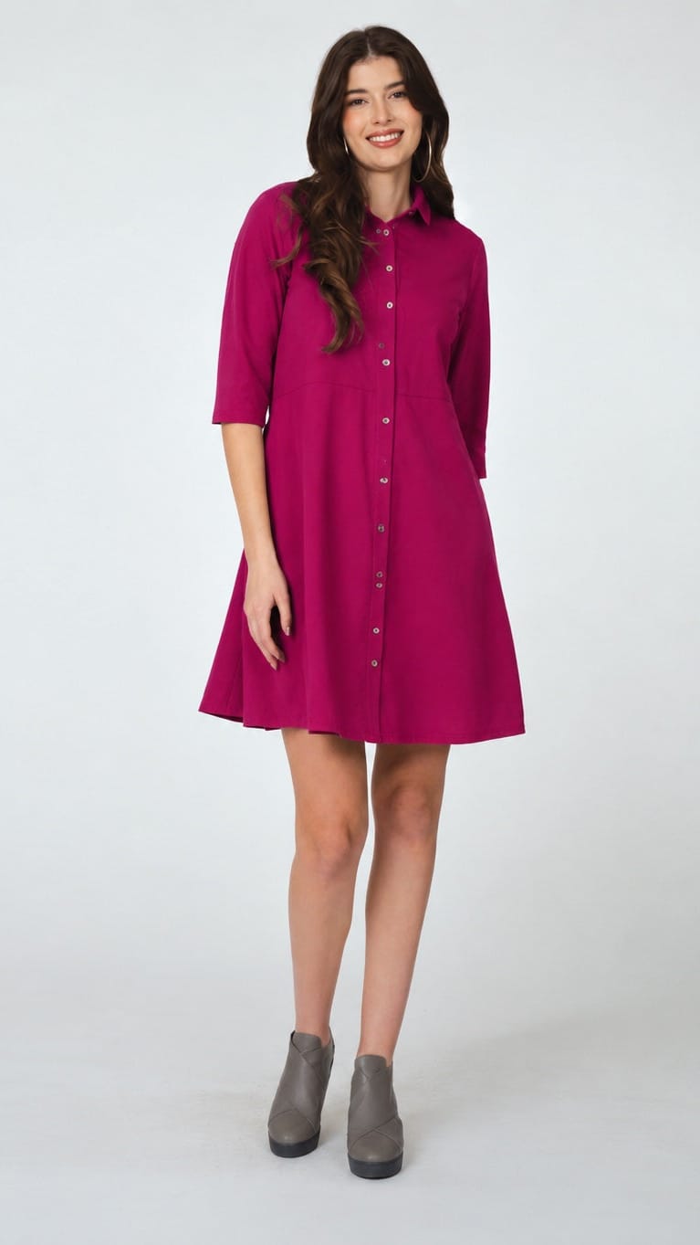A woman modeling a magenta pink button-down shirt dress with three-quarter sleeves and a flared skirt, paired with gray ankle boots, against a light gray studio background.