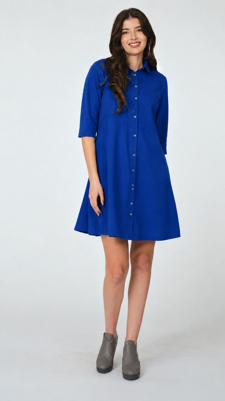 A woman modeling a bright royal blue button-down shirt dress with three-quarter sleeves and a flared skirt, paired with gray ankle boots, against a light gray studio background.