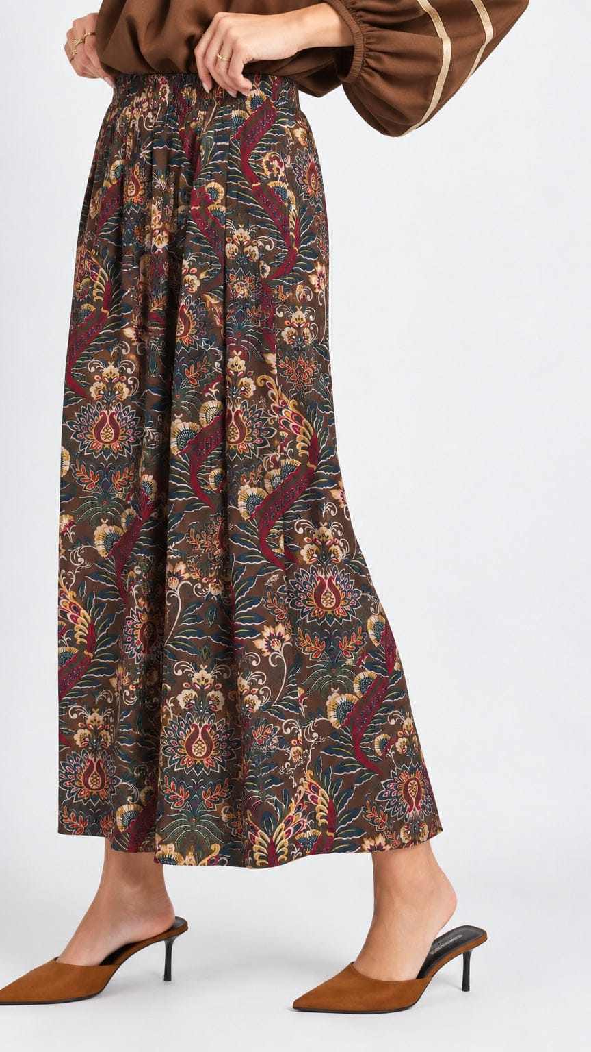 A model wearing a brown and multicolor paisley patterned maxi skirt with a brown top and brown mule heels, shown from the waist down against a light gray studio background.