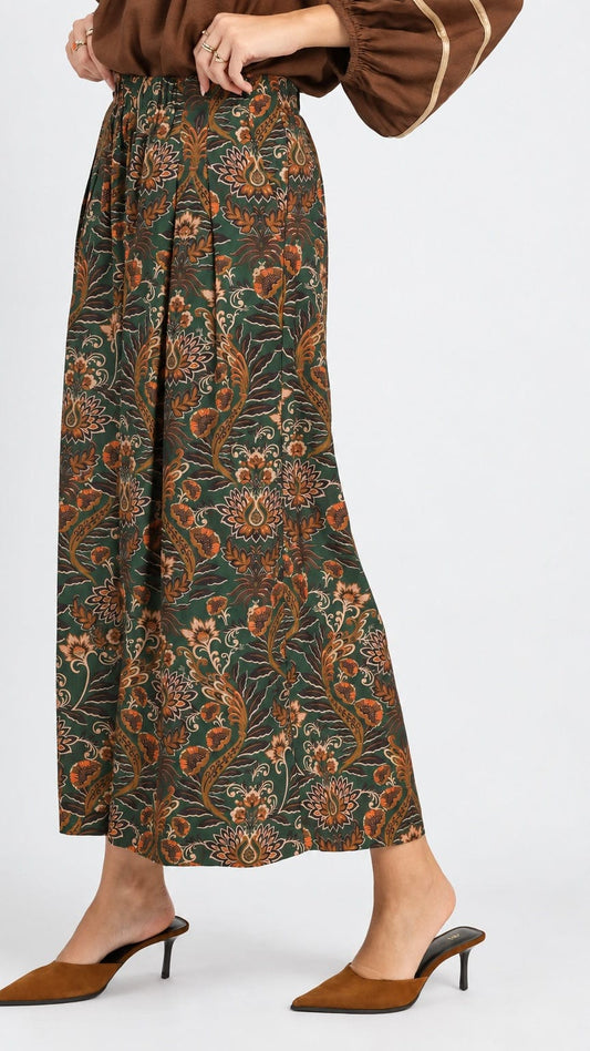 A model wearing a pair of dark green wide-leg trousers with an intricate orange and brown floral pattern, paired with brown mule heels against a light gray studio background.