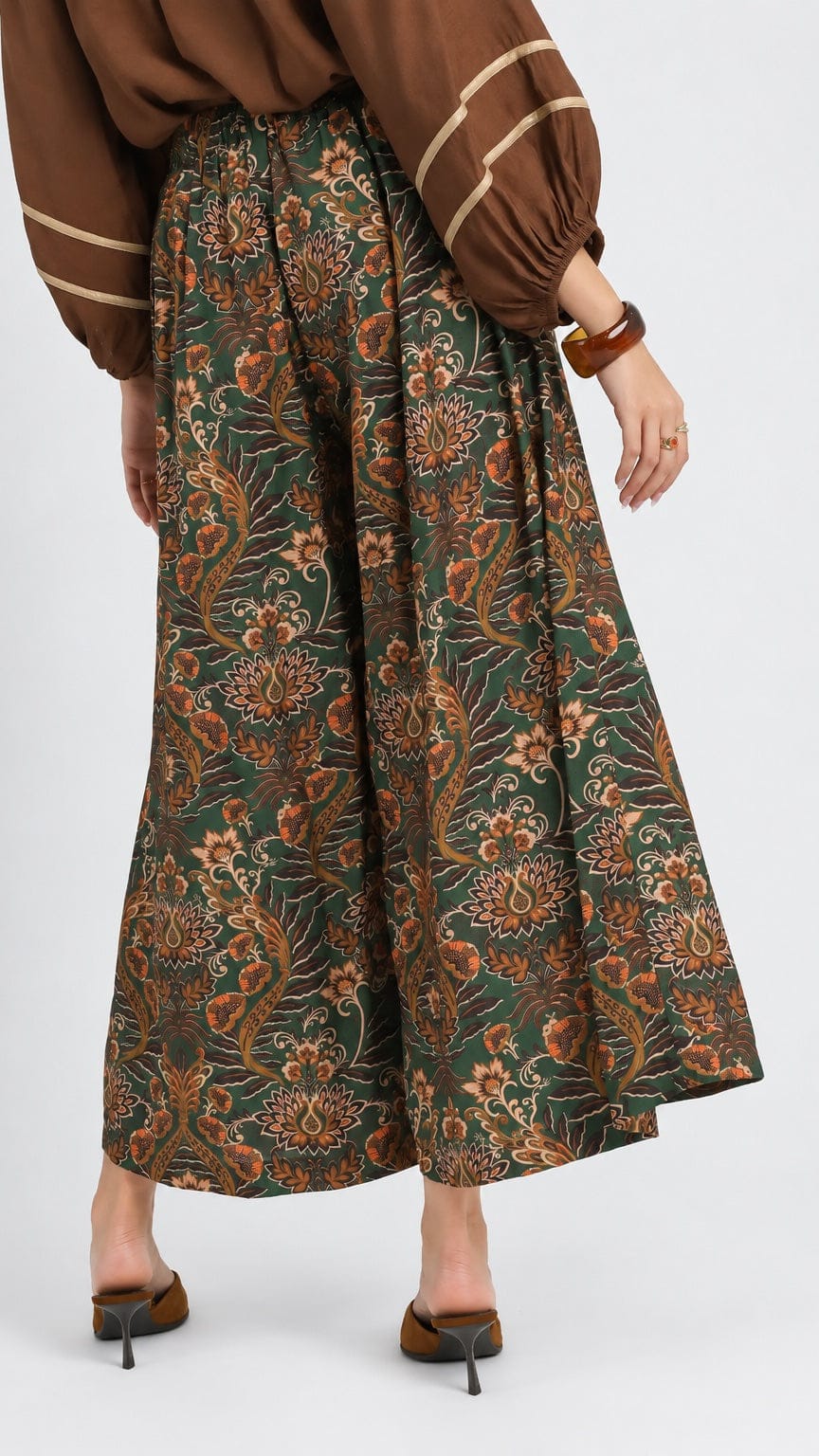 A model from the back wearing a brown and green floral patterned wide-leg pleated skirt, paired with a brown blouse and brown heels, against a light gray studio background.