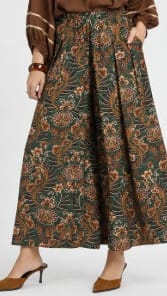 A model wearing a brown and green floral patterned pleated maxi skirt with a matching brown top and brown pointed-toe heels, shown from the waist down on a light gray background.