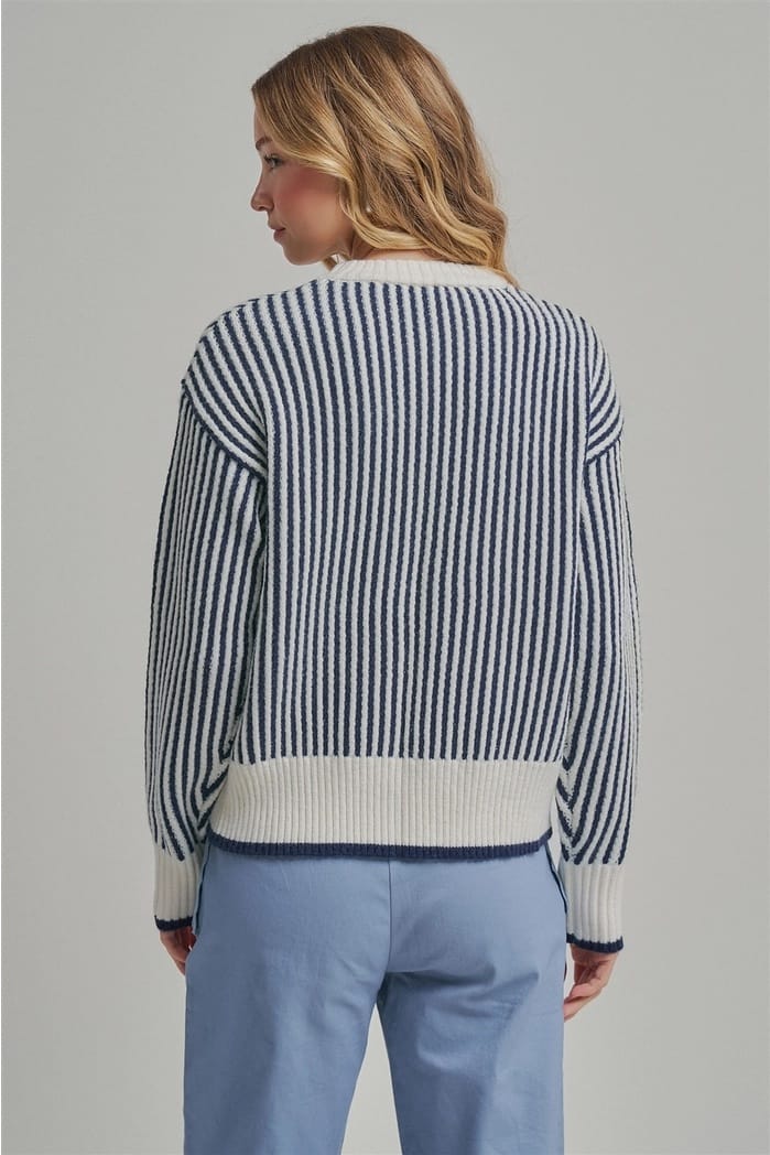 A woman shown from the back wearing a navy and white vertically striped knit sweater with solid white ribbed trim, paired with light blue trousers, against a solid gray studio background.