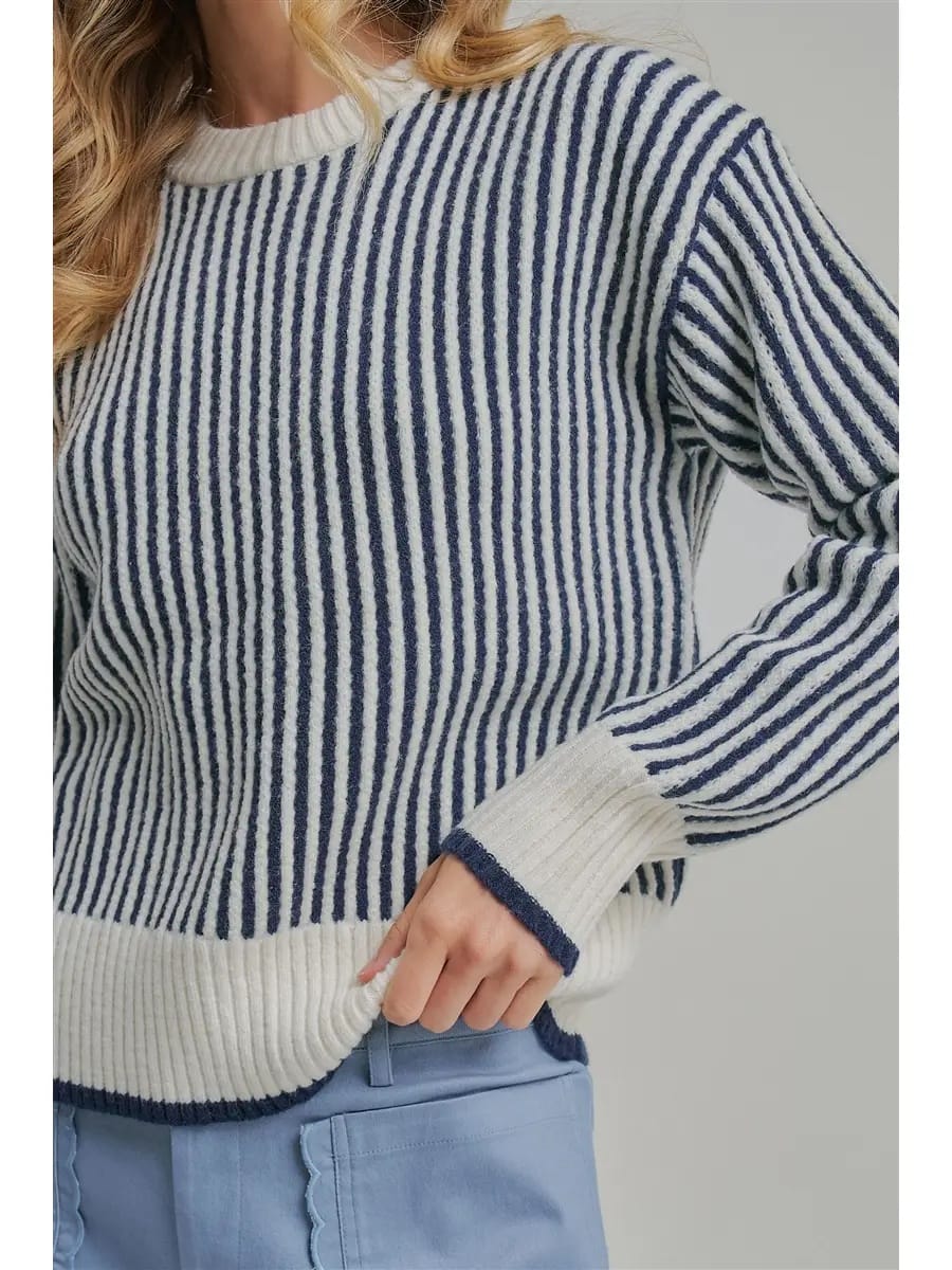 A close-up of a woman wearing a ribbed knit sweater with vertical navy and off-white stripes, paired with light blue pants, against a neutral gray background.