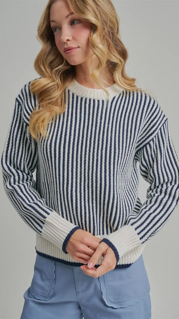 A woman wearing a navy and cream vertically striped knit sweater with ribbed cuffs and hem, paired with light blue jeans, against a solid gray studio background.