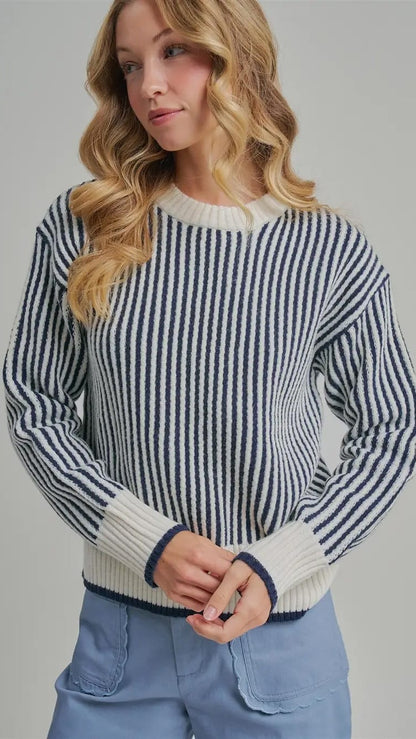 A woman wearing a navy and cream vertically striped knit sweater with ribbed cuffs and hem, paired with light blue jeans, against a solid gray studio background.