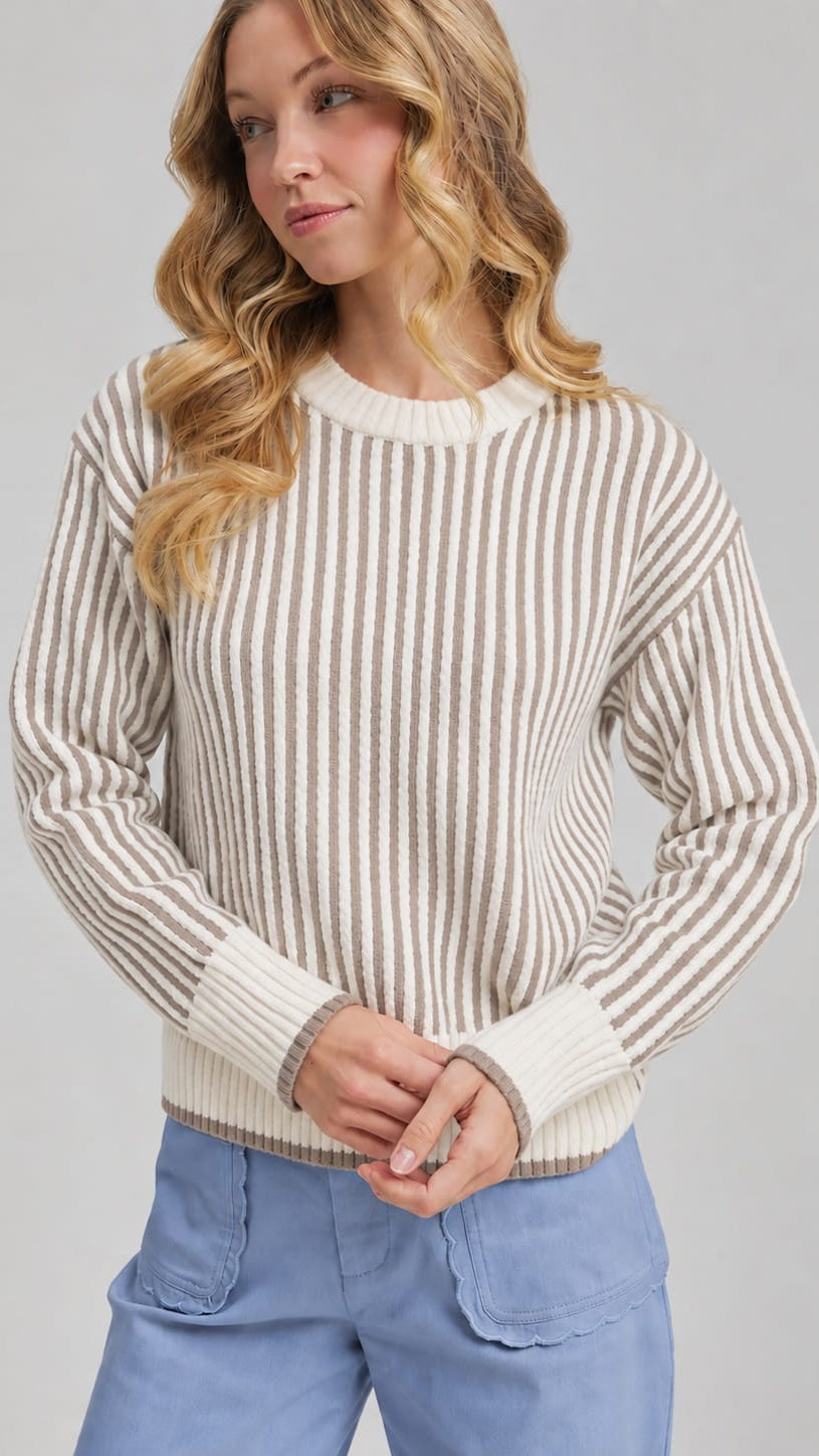 A woman wearing a cream and brown vertically striped knit sweater with light blue pants, photographed against a plain light gray studio background.