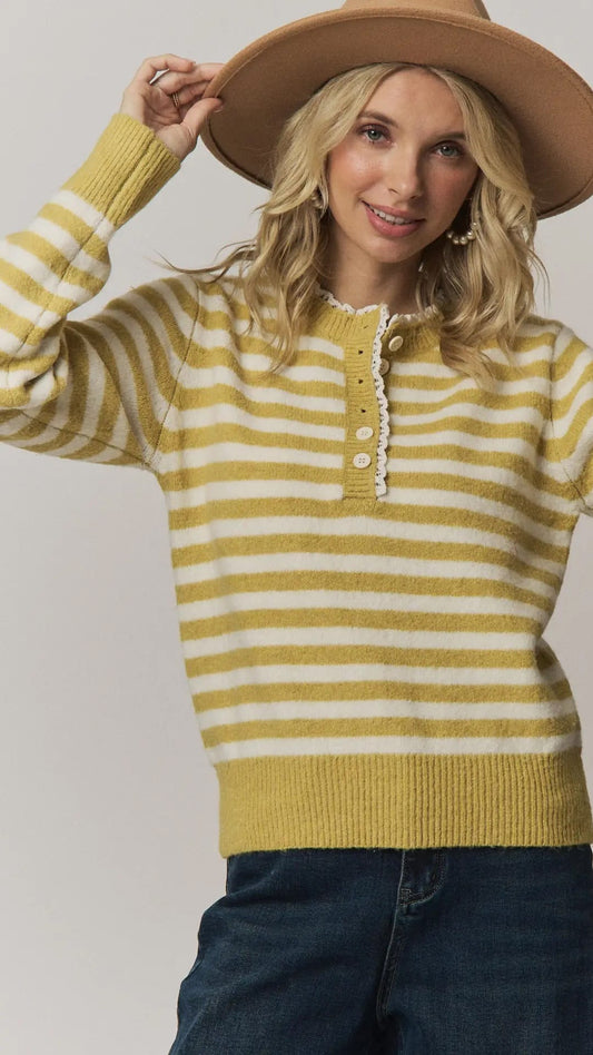 A woman wearing a yellow and white striped knit henley sweater, a wide-brim tan hat, and dark blue jeans, posing against a light gray studio background.
