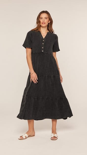 A woman modeling a short-sleeve black tiered midi dress with a buttoned V-neckline, photographed against a plain light beige studio background.
