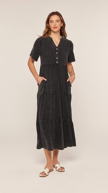 A woman modeling a dark charcoal gray short-sleeve midi dress with a tiered skirt, buttoned V-neckline, and side pockets, paired with white slide sandals, against a plain light beige studio background.
