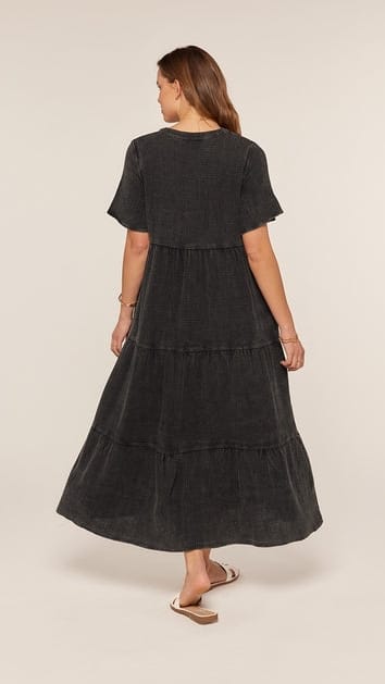 A woman wearing a dark charcoal gray tiered midi dress with short sleeves, shown from the back against a plain beige studio background.
