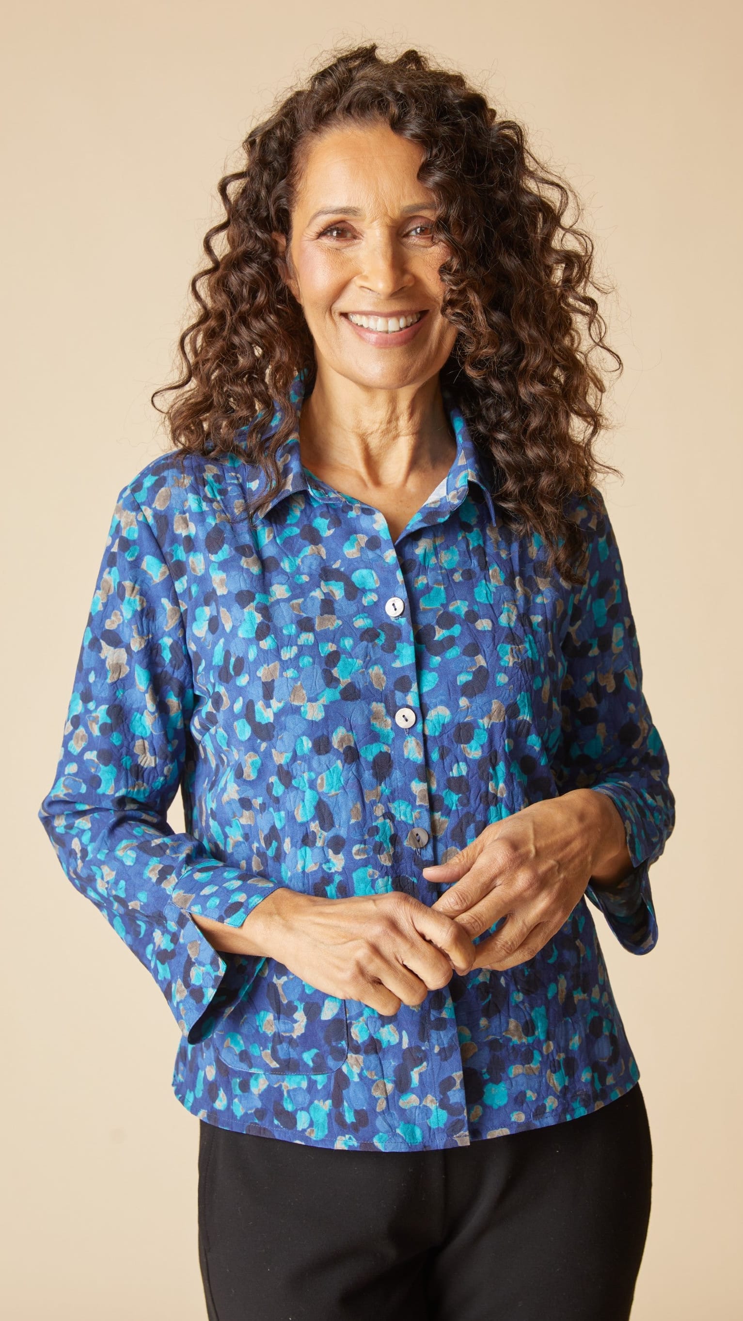 A smiling woman with long curly brown hair wearing a blue button-up shirt with a multicolor abstract pattern, paired with black pants, against a solid beige background.