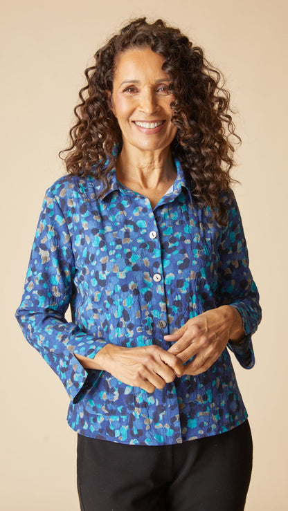 A smiling woman with long curly brown hair wearing a blue button-up shirt with a multicolor abstract pattern, paired with black pants, against a solid beige background.