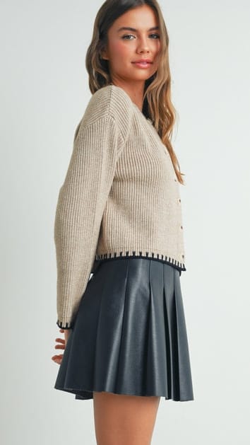 A woman wearing a beige ribbed knit cardigan with black pom-pom trim and a dark gray pleated mini skirt, shown from a side-back angle against a light gray studio background.