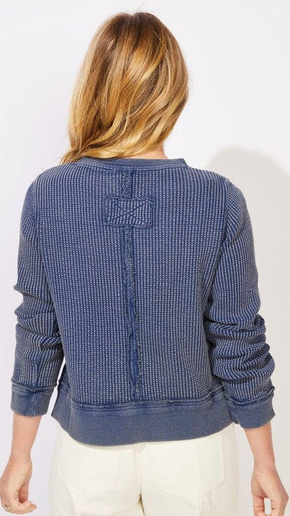 A woman viewed from the back wearing a textured blue knit sweater with cream pants, set against a plain white studio background.