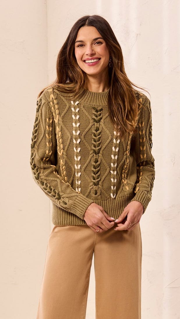 A woman stands indoors wearing a textured olive green cable-knit sweater with cream leaf patterns, tan wide-leg cropped pants, and brown ankle boots against a soft cream curtain backdrop.