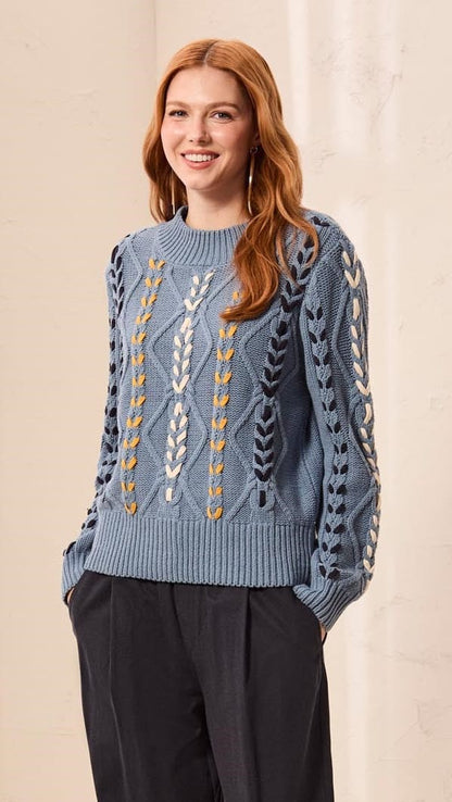 A woman modeling a light blue knit sweater with intricate white, yellow, and gray geometric patterns on the front and sleeves, paired with dark charcoal wide-leg trousers and brown suede loafers, set against a warm cream-colored wall and floor.