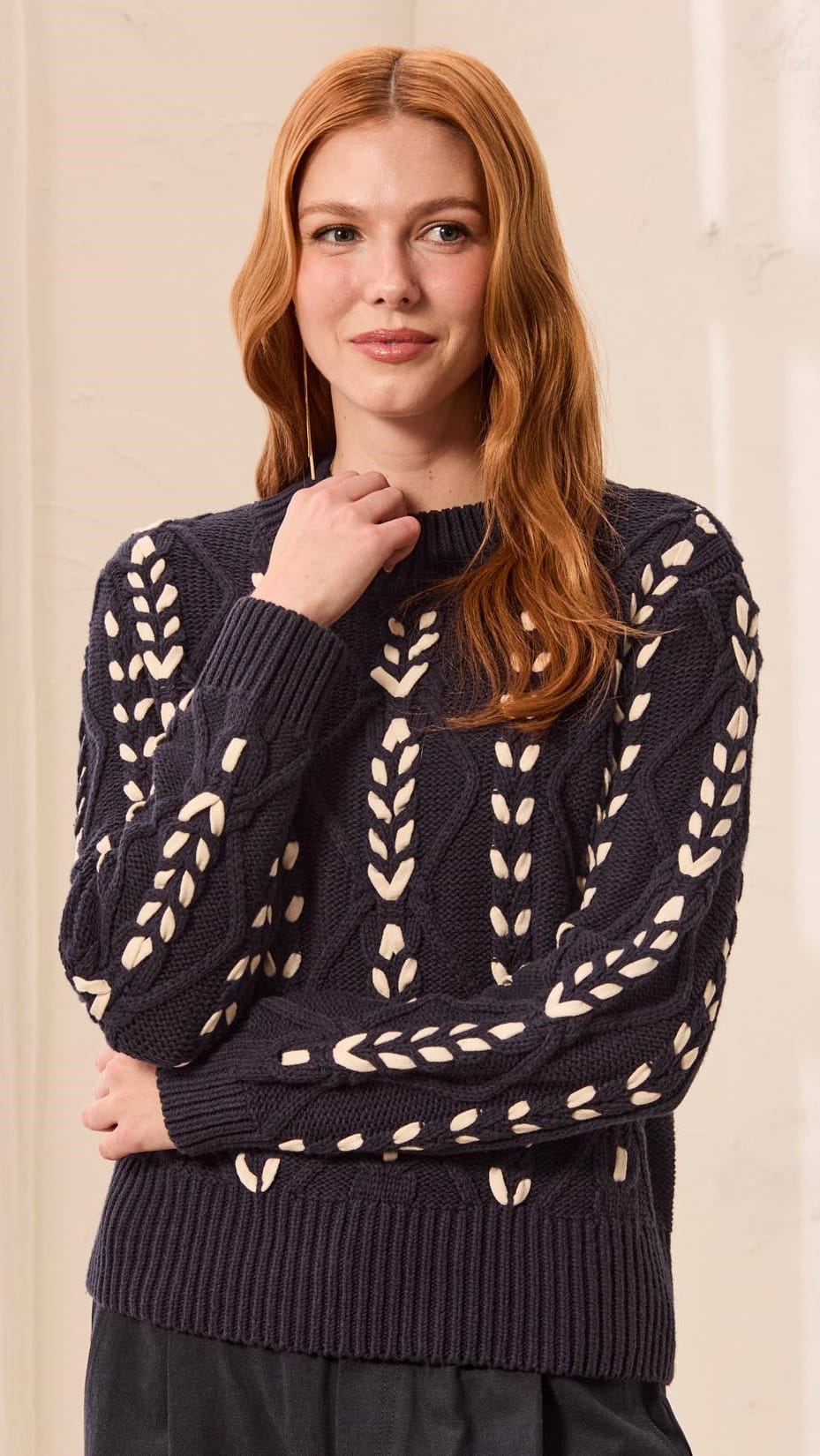 A woman with long red hair wearing a dark navy cable-knit sweater with cream leaf patterns, posed against a soft beige background.