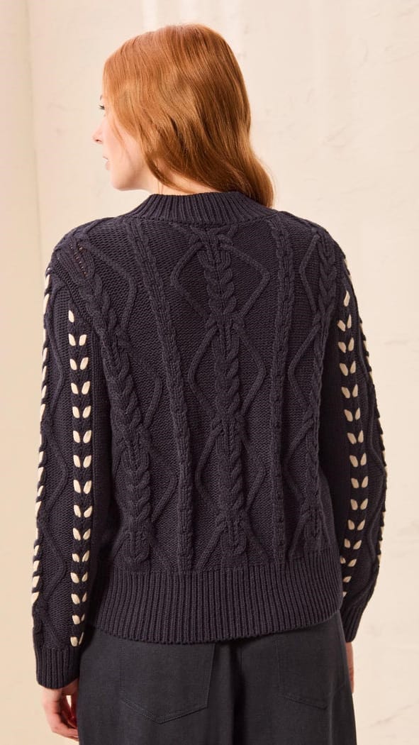 A woman with red hair is shown from the back wearing a dark navy cable-knit sweater with decorative cutout details on the sleeves, paired with dark trousers.