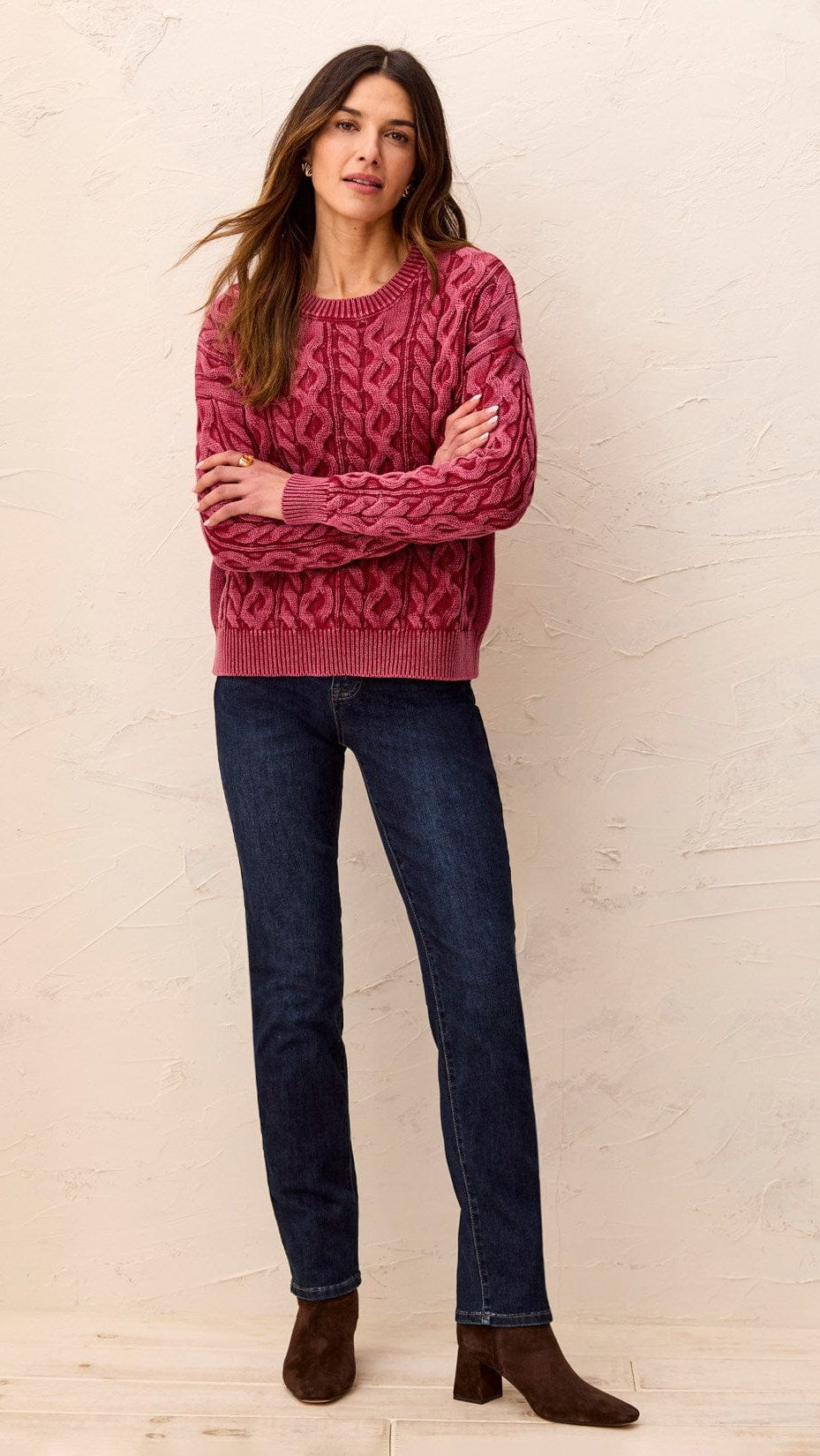A woman modeling a dusty rose cable-knit sweater, dark blue jeans, and brown ankle boots, standing against a textured beige wall.