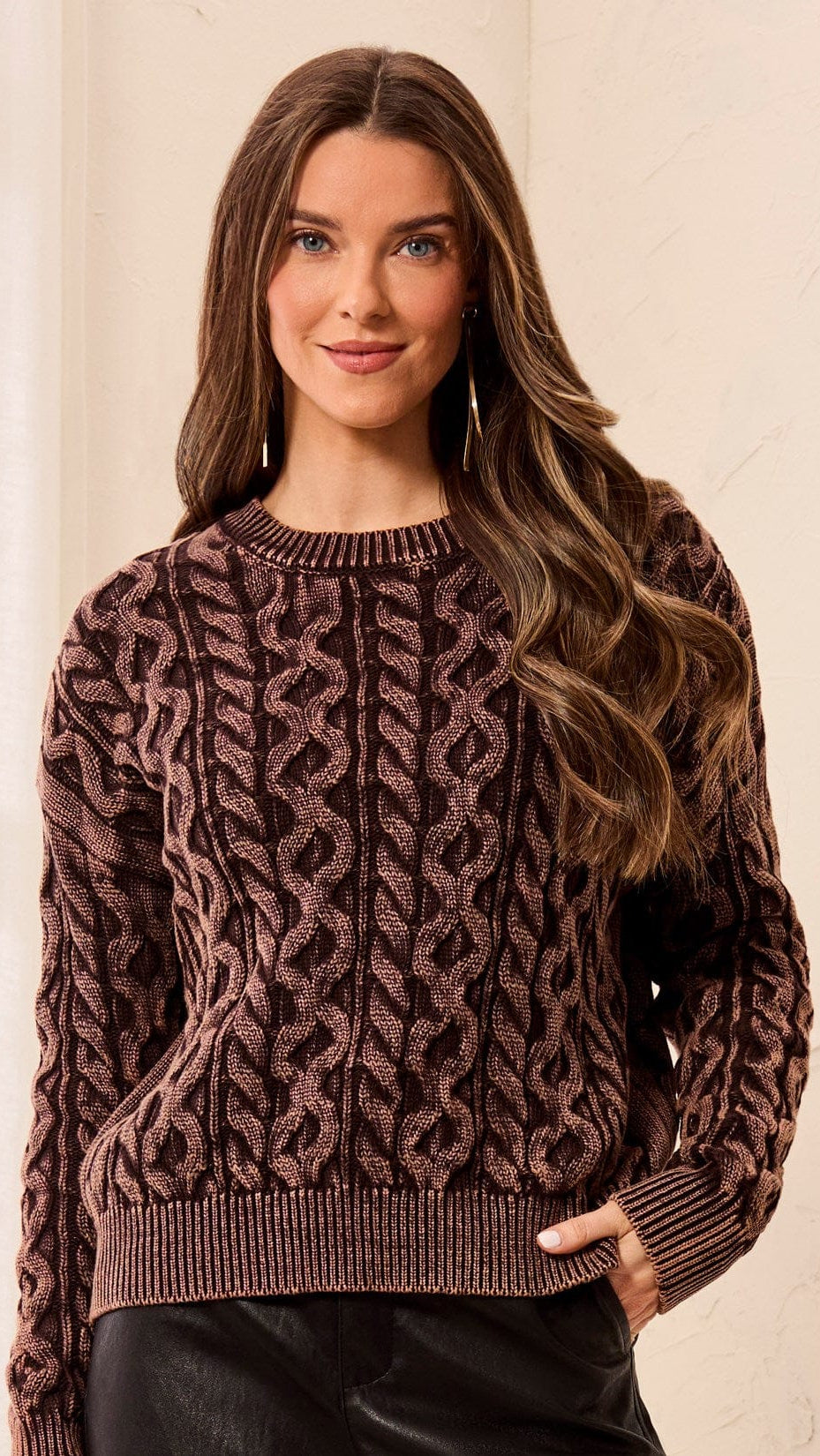 A woman wearing a brown and black cable-knit crewneck sweater, standing against a light beige textured wall.