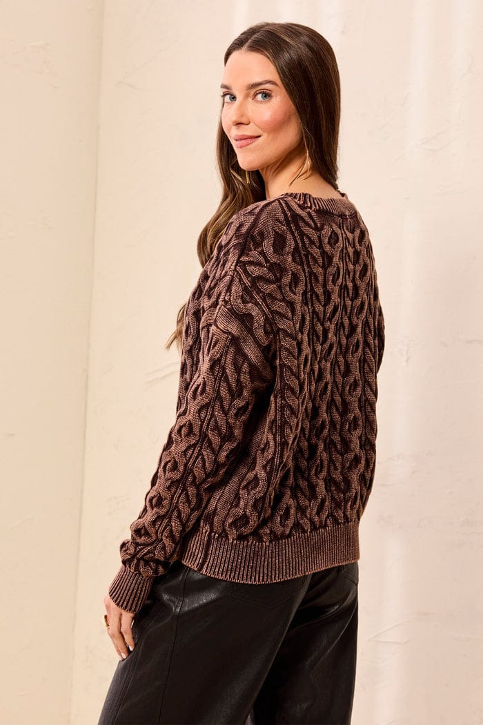 A woman wearing a brown cable-knit sweater with a textured pattern, shown from a side-back angle against a light beige wall.