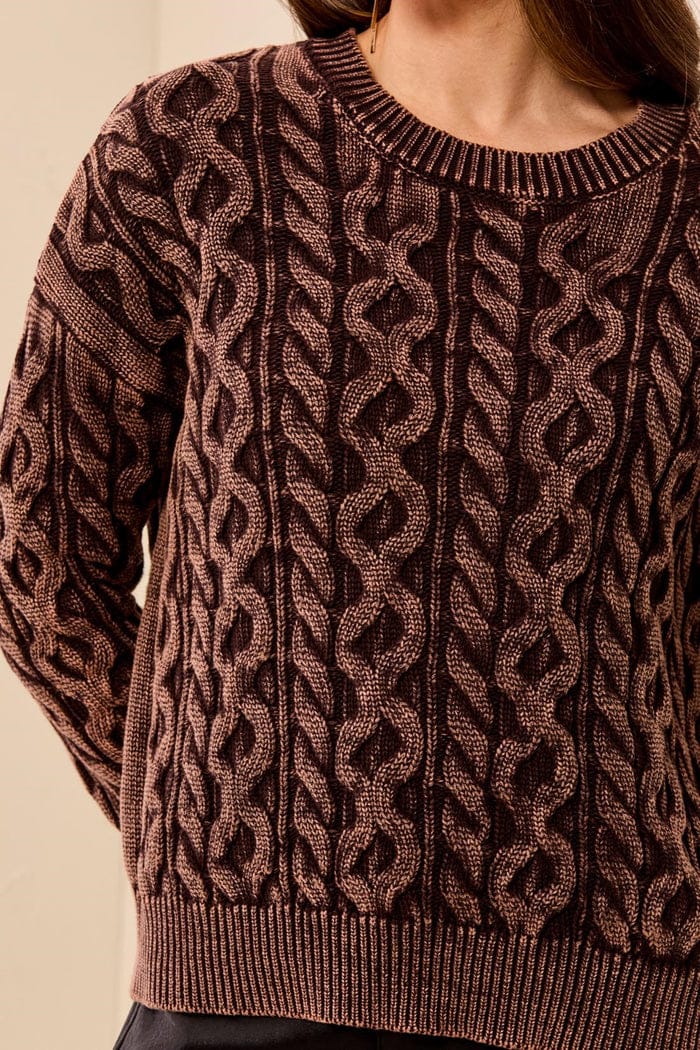 A close-up of a person wearing a textured cable-knit sweater in a dark brown and muted pinkish-brown colorway, showing the detailed knit pattern and ribbed collar, cuffs, and hem.