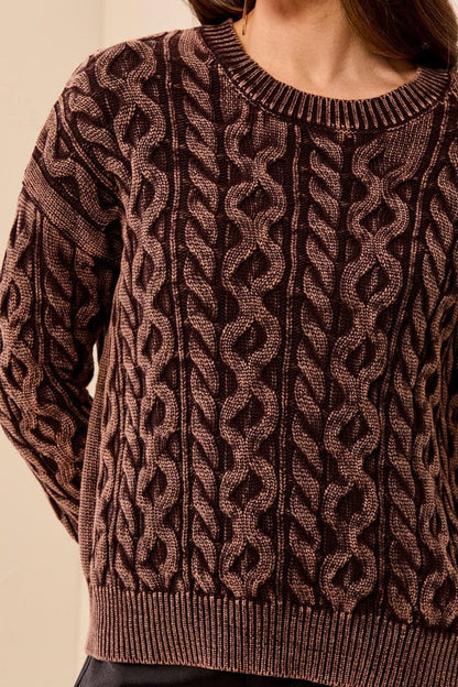 A close-up of a person wearing a textured cable-knit sweater in a dark brown and muted pinkish-brown colorway, showing the detailed knit pattern and ribbed collar, cuffs, and hem.
