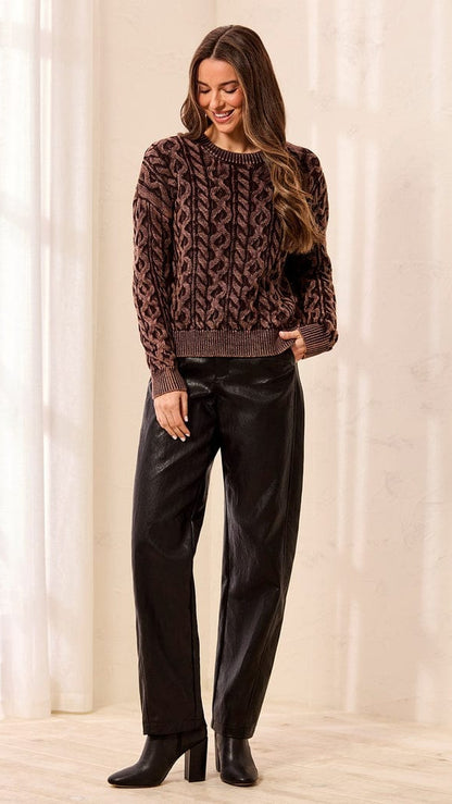 A woman modeling a brown and black patterned knit sweater paired with black pants and black ankle boots, standing in a brightly lit indoor setting with soft curtains.