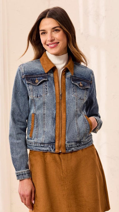 A woman wearing a blue denim jacket with a brown collar and zipper trim, layered over a white turtleneck and a brown skirt, set against a soft beige background.