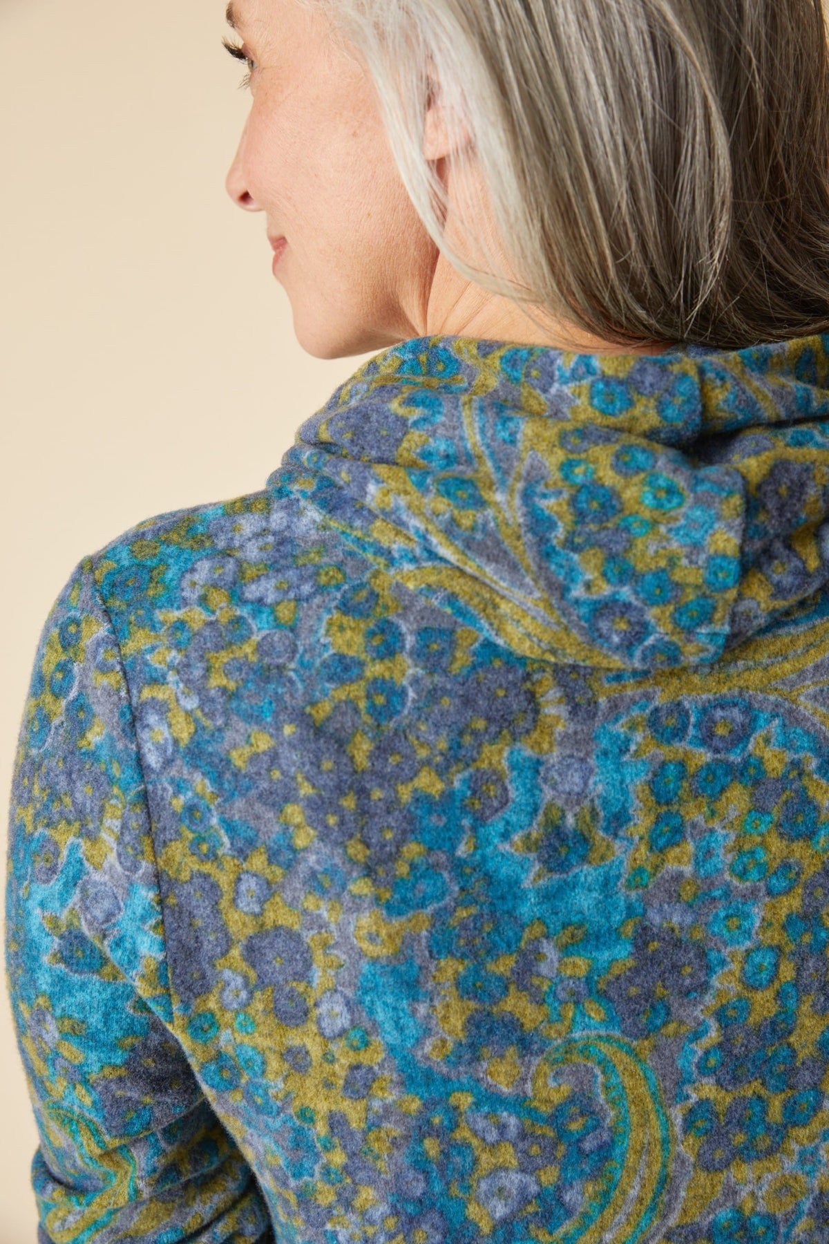 A close-up side profile of a woman wearing a hooded sweatshirt with a textured paisley pattern in blue, teal, and olive green, set against a plain beige background.